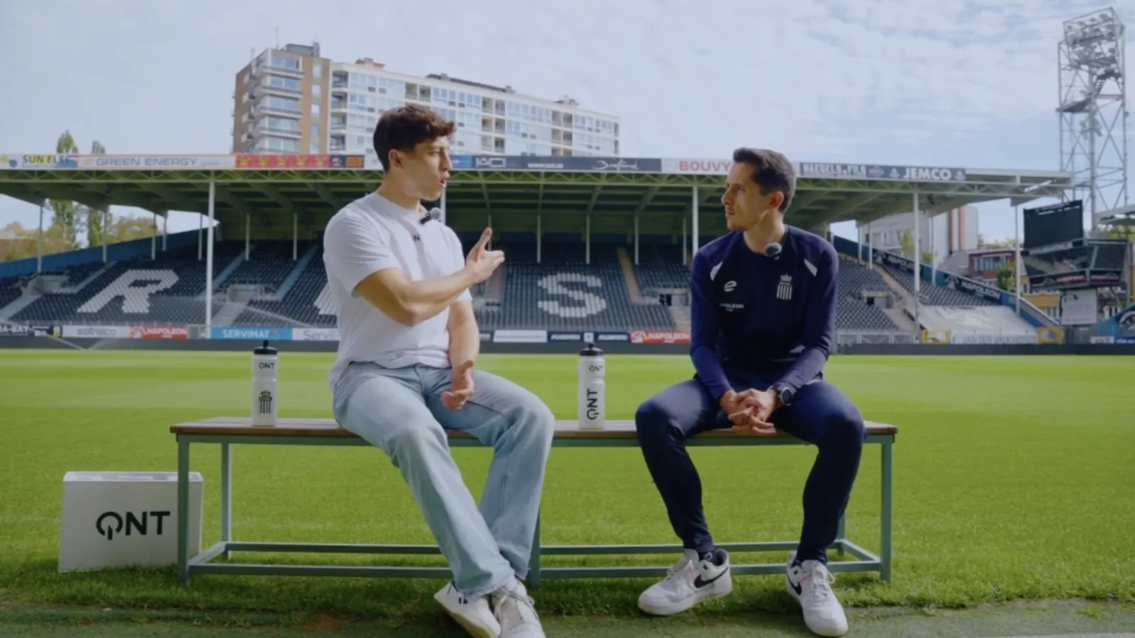 Marius Grek, assis sur un banc au bord d'un terrain de football, mène un entretien avec un invité en tenue de club, devant une tribune vide